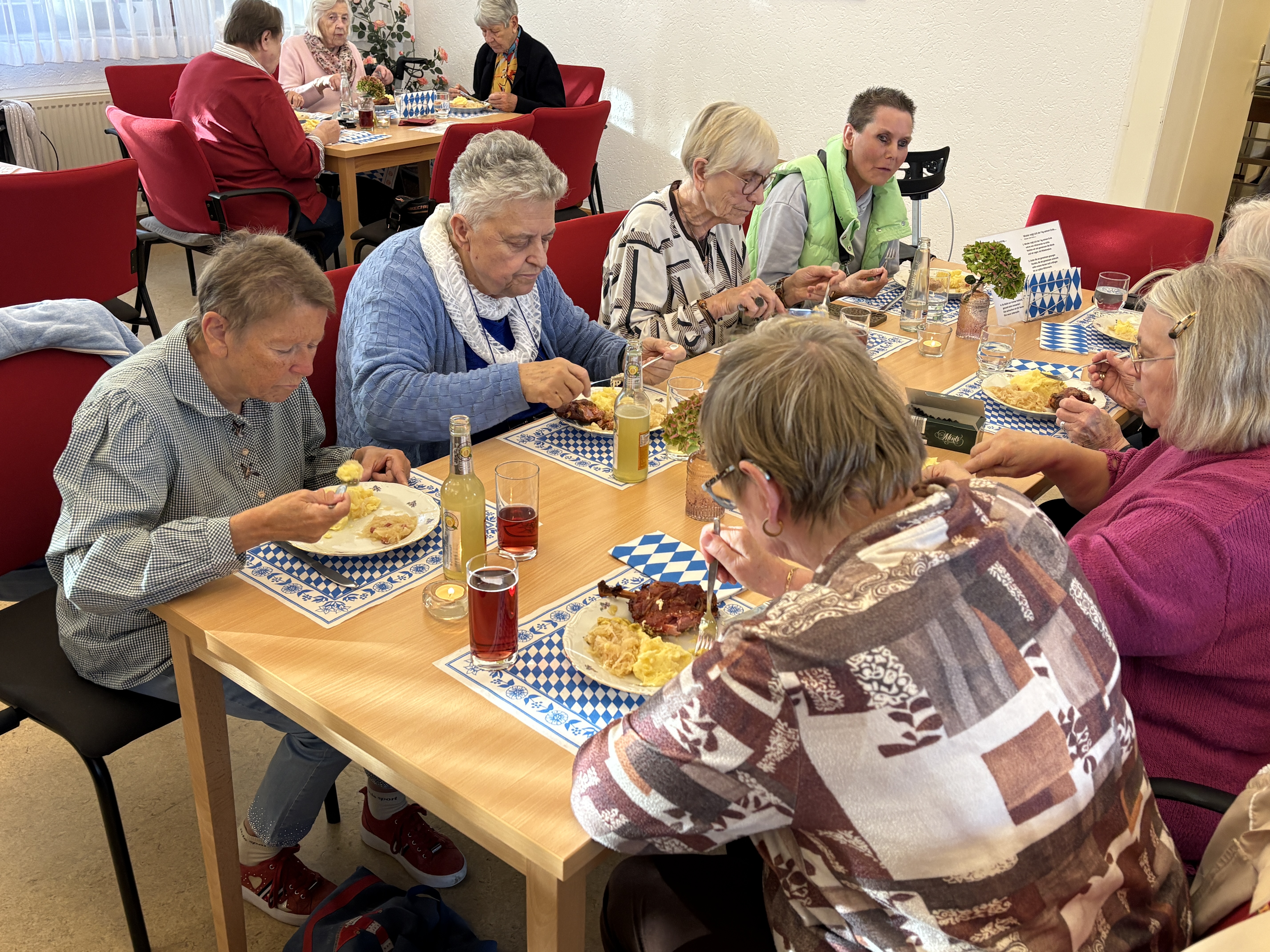 Seniorinnen sitzen an Tischen mit blau-weißen Platzsets und essen Haxe mit Kartoffeln