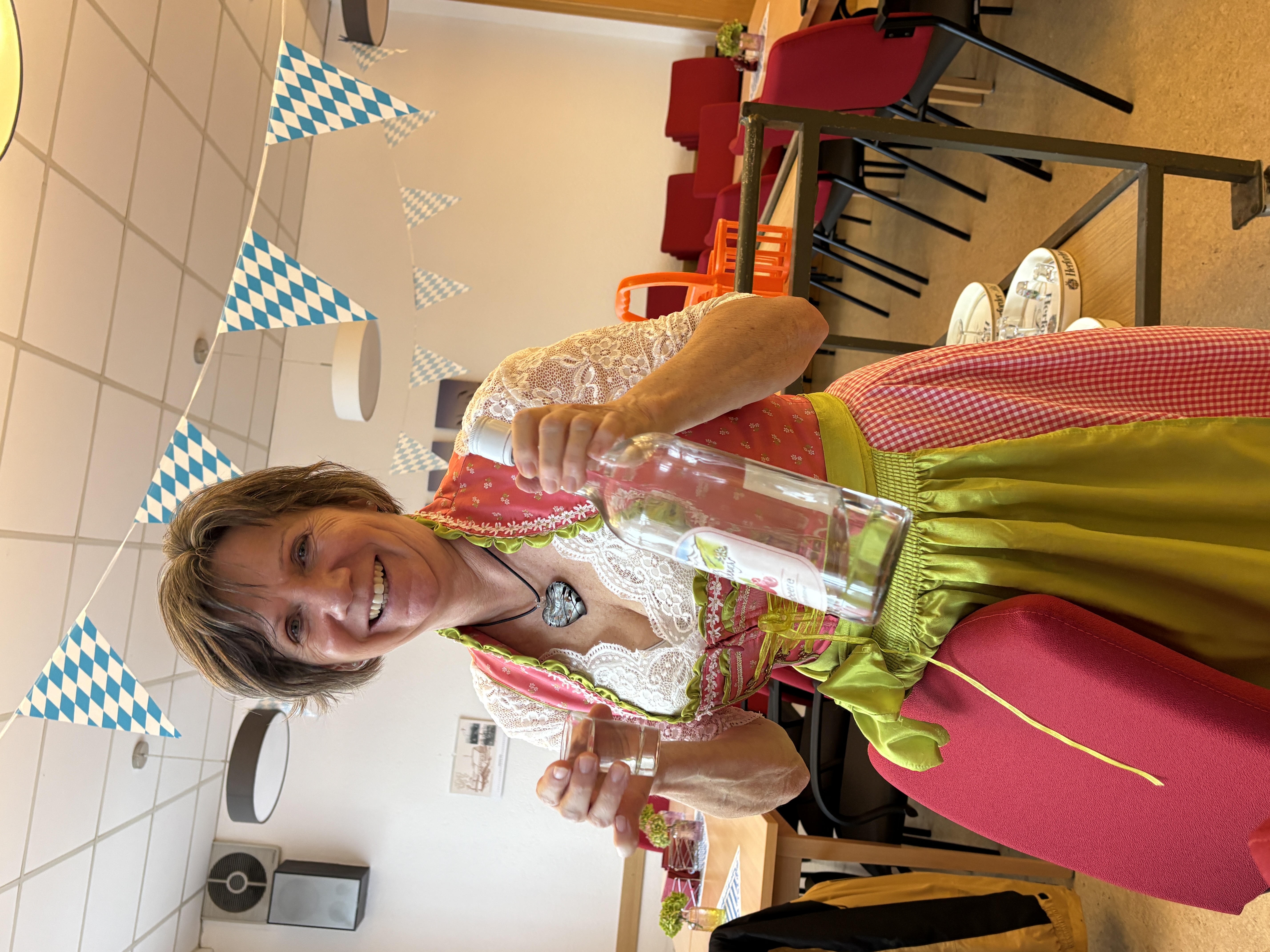 Frau im Dirndl hält lachend eine Flasche und ein Glas unter blau-weißen Wimpeln