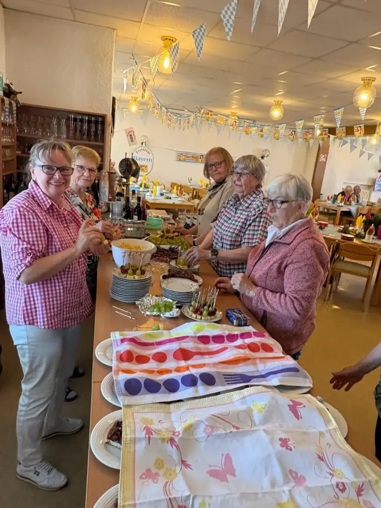 Frauen stehen lächelnd an einem langen Buffet mit Spießen, Salat und Tellern, im Hintergrund der geschmückte Saal