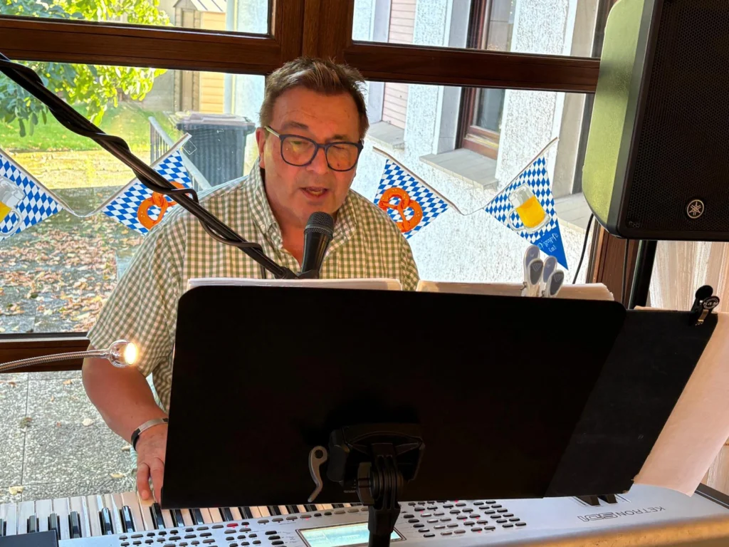 Mann in kariertem Hemd spielt Keyboard und singt ins Mikrofon, hinter ihm blau-weiße Wimpel am Fenster
