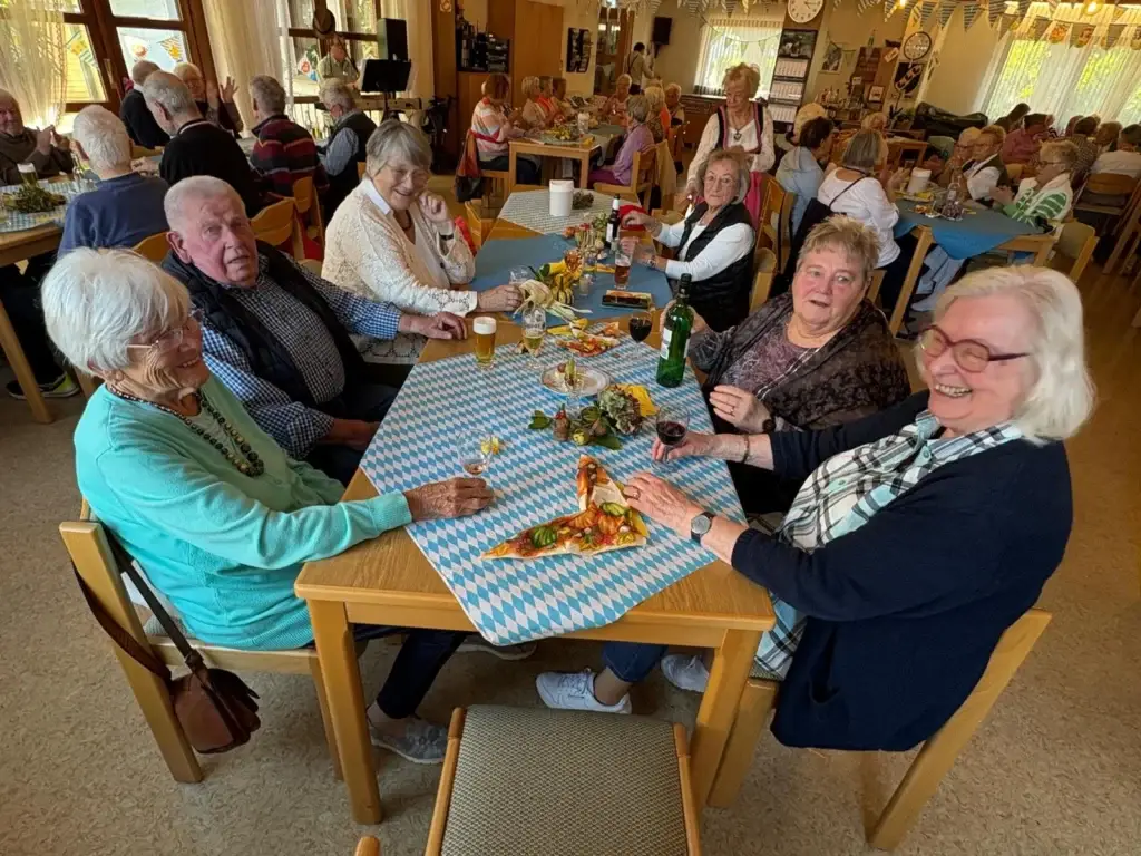 Seniorinnen und Senioren sitzen fröhlich an einem Tisch mit blau-weiß gerauteter Tischdecke, Bier und Wein, dahinter der voll besetzte Saal
