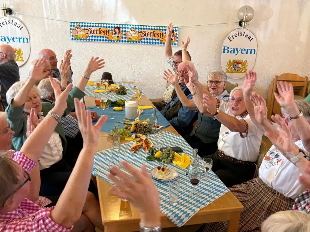 Gäste an einer langen Tafel unter Schildern „Freistaat Bayern“ und einem Bierfest-Banner recken die Hände in die Höhe