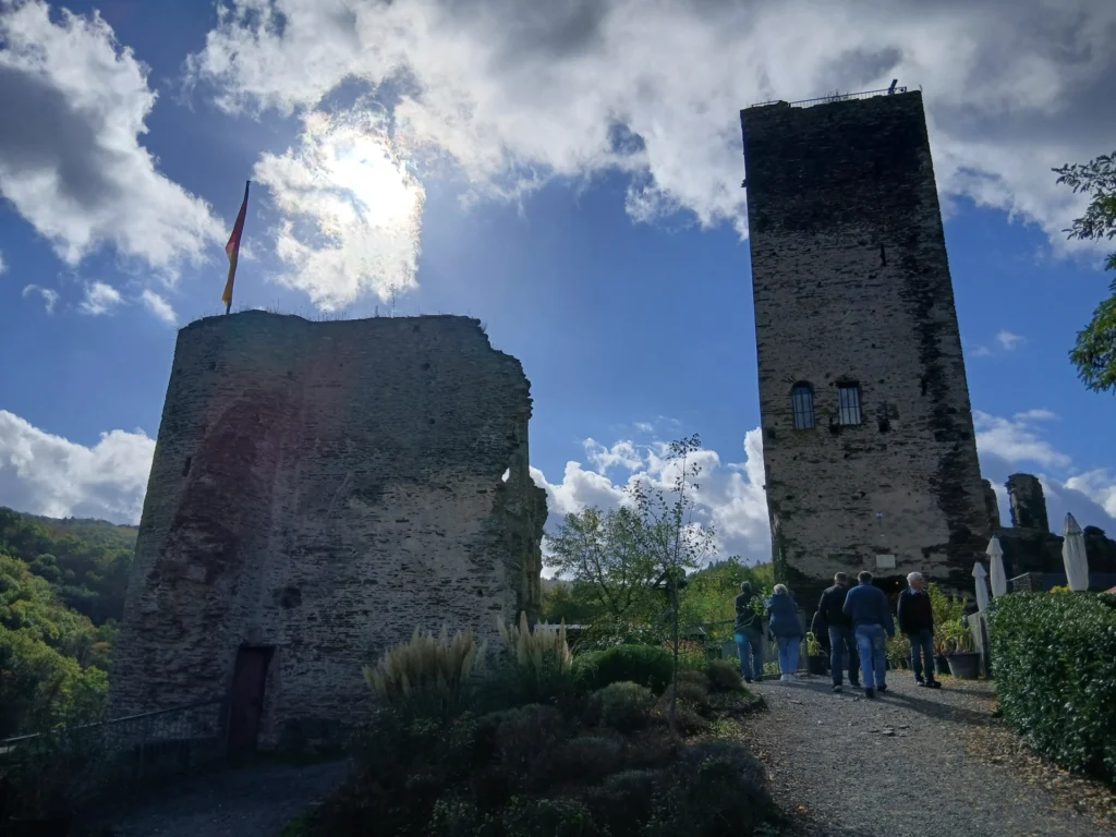 Der hohe Bergfried der Burgruine Metternich und ein weiterer Mauerturm mit Fahne vor blauem Himmel, Besucher gehen den Weg entlang