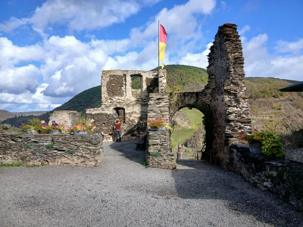 Mauerreste und ein Torbogen aus Bruchstein in der Burgruine Metternich, mit Blumenkübeln und einer Fahne, im Hintergrund bewaldete Hänge