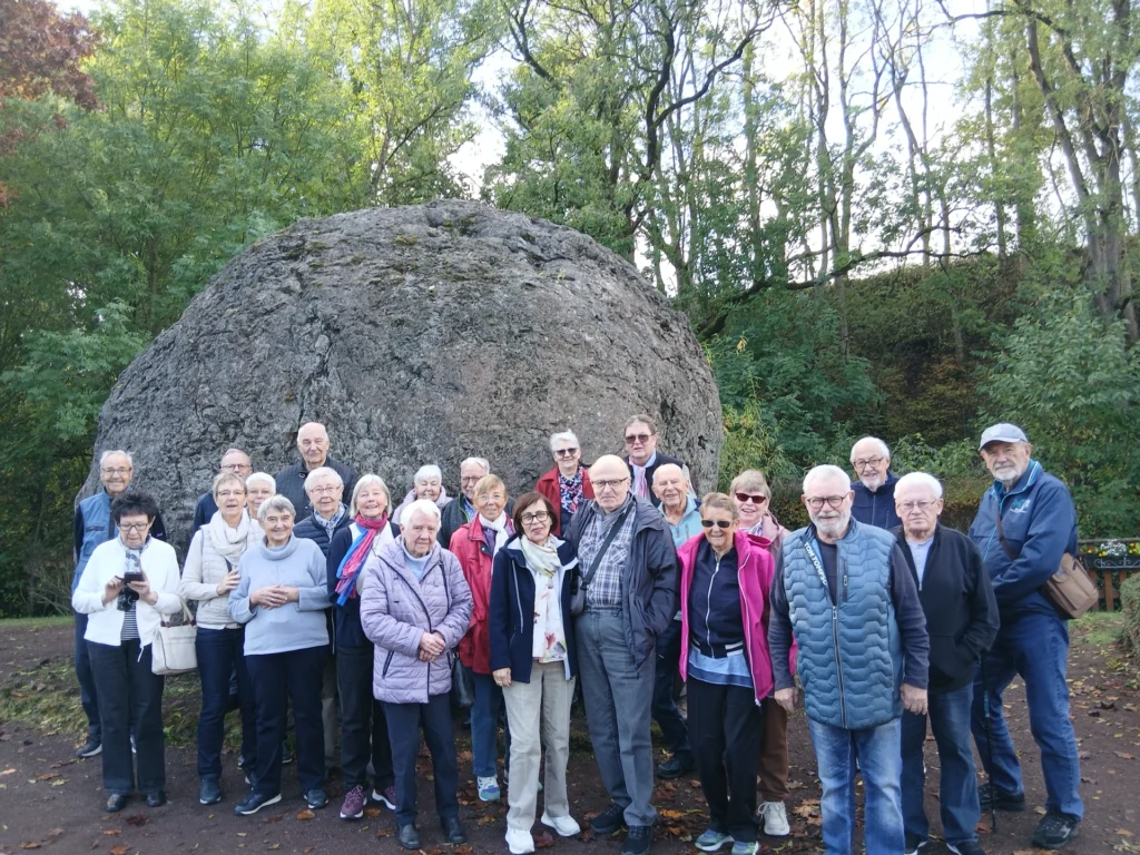 Die Reisegruppe steht vor einem riesigen, runden Gesteinsbrocken, der Lavabombe von Strohn, umgeben von Bäumen