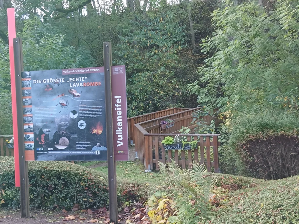 Infotafel des Vulkan-Erlebnispfads Strohn mit der Überschrift „Die größte ‚echte‘ Lavabombe“, dahinter ein Holzsteg zwischen Bäumen