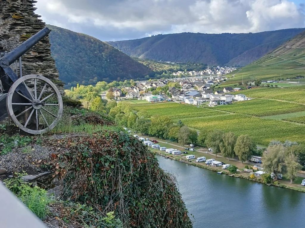 Eine alte Kanone mit Holzrad an der Burgmauer, dahinter die Mosel mit einem Campingplatz am Ufer, Weinberge und der Ort Ellenz-Poltersdorf
